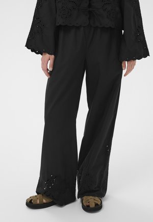 Personne portant un pantalon noir à jambes larges avec des détails en dentelle ajourée aux ourlets et des sandales en daim marron avec des semelles noires, debout sur un fond blanc.