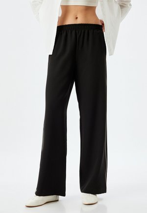 Zwarte wijde broek met een elastische tailleband, gemaakt van gladde stof. Bevat zijsplitten en twee verticale witte strepen langs de naden.