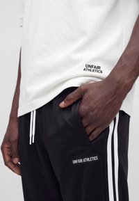 Muž nosiaci biele polo tričko a čierne športové šortky s bielymi pruhmi, oboje značky "UNFAIR ATHLETICS", s jednou rukou v vrecku šortiek.