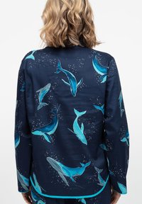 Camicia blu navy con motivo di balene in turchese. Realizzata in tessuto liscio, ha maniche lunghe e un orlo curvo con un bordo in contrasto.
