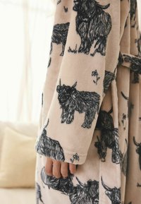 Puha bézs köntös fekete vázlatokkal, melyeken bolyhos highlandi tehenek láthatók, a fél kéz az ujjszegélynél részben látható természetes beltéri fényben.