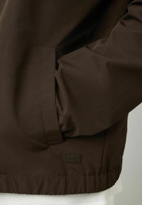Veste légère marron avec une texture lisse, dotée de poches latérales et d'un ourlet côtelé. Comprend un petit logo de la marque dans le coin inférieur.