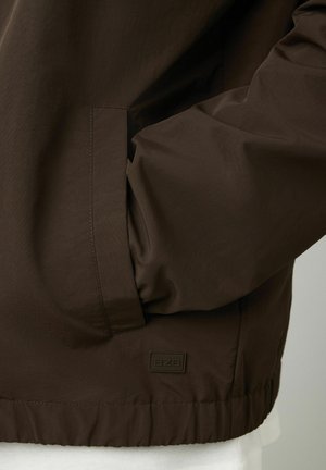 Veste légère marron avec une texture lisse, dotée de poches latérales et d'un ourlet côtelé. Comprend un petit logo de la marque dans le coin inférieur.