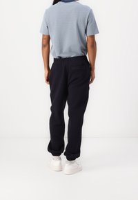 Man som bär en ljusblå kortärmad skjorta, svarta joggerbyxor med elastiska mudd, vita sneakers, står vänd mot en vit vägg.