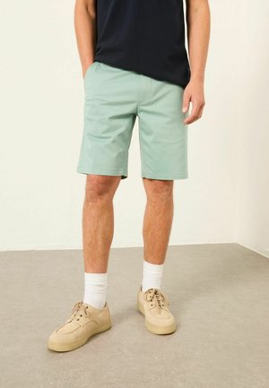 Mann trägt mintgrüne Shorts, beige Sneakers mit weißen Kniestrümpfen und ein schwarzes T-Shirt, steht auf einem grauen Boden vor einer weißen Wand.