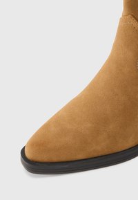 Barna velúr bokacsizma hegyes orral és fekete gumitalppal. Simaságot és minimális varrást kínál a letisztult megjelenés érdekében.