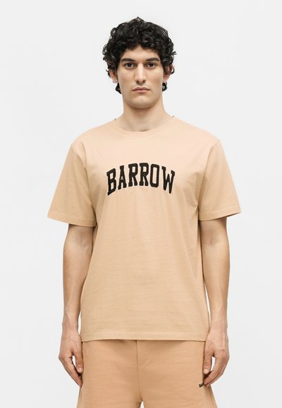 Άνδρας με σγουρά σκούρα μαλλιά, που φοράει μπεζ μπλουζάκι και ταιριαστό σορτς με τη λέξη «BARROW» στο μπροστινό μέρος.
