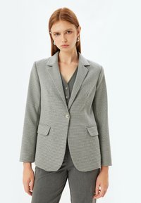 Harmaa houndstooth-takki, jossa on ärhäkkä kaulus, yksittäinen nappisulku, kaksi etutaskua ja pitkät hihat, valmistettu  tekstuurista kankaasta.