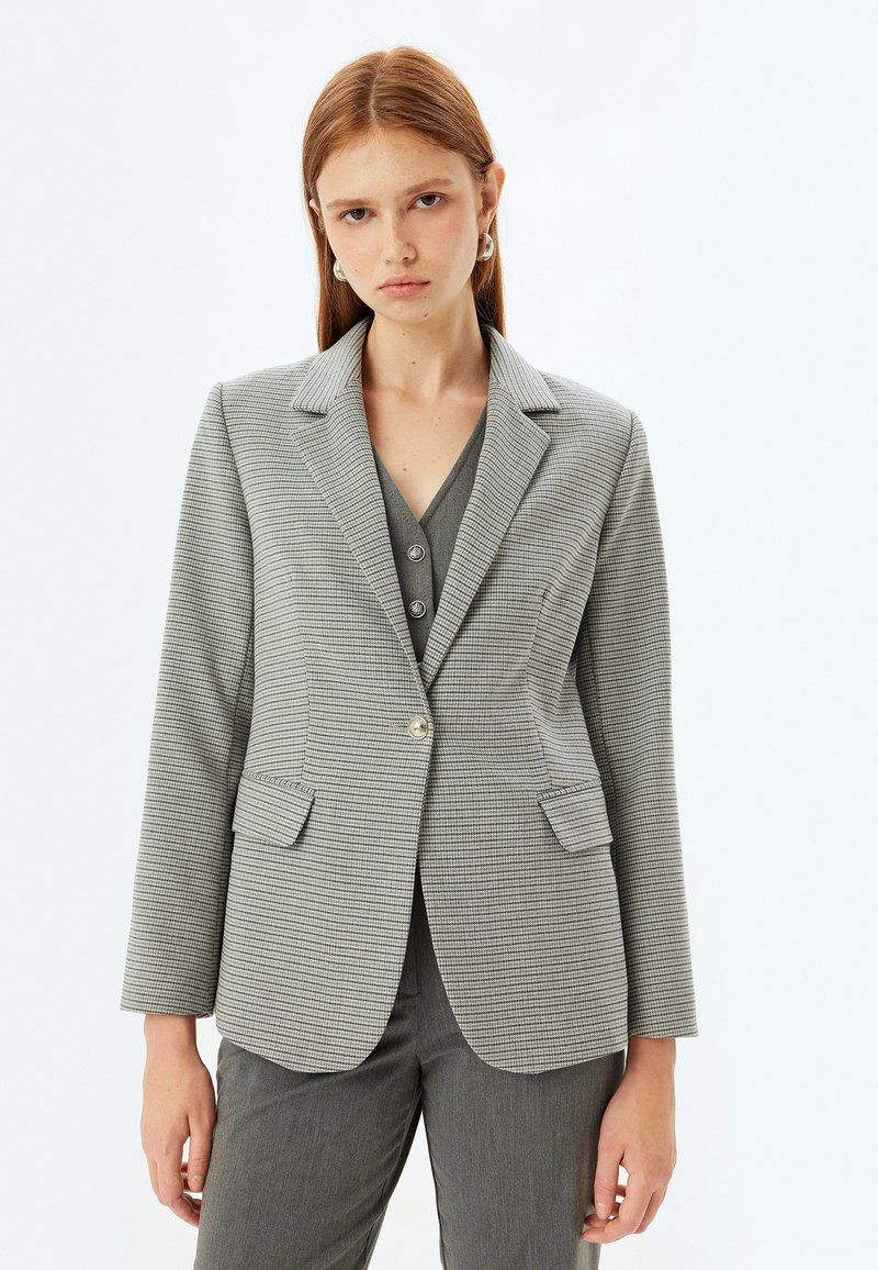 Harmaa houndstooth-takki, jossa on ärhäkkä kaulus, yksittäinen nappisulku, kaksi etutaskua ja pitkät hihat, valmistettu  tekstuurista kankaasta.