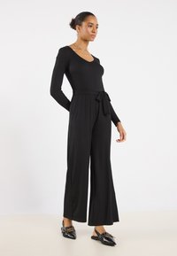 Black jumpsuit with long sleeves, V-neck, belted waist, and wide-leg trousers. Smooth texture with a fitted top, elegant design.