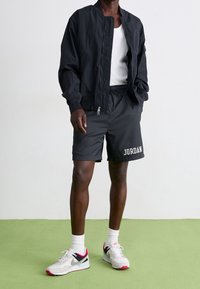 Muž ve černé bundě na zip, bílém tílku, černých šortkách s nápisem "JORDAN", bílých ponožkách a bílých teniskách Nike s růžovými akcenty.