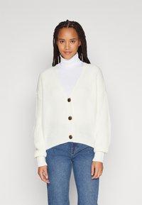White, textured knit cardigan with a V-neck, five buttons, and balloon sleeves, paired with blue jeans.