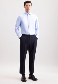 Man staat met zijn handen in zijn zakken, gekleed in een lichtblauw overhemd met knopen, zwarte broek en zwarte nette schoenen, tegen een effen achtergrond.