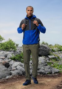 Mėlyna ir pilka zip-up striukė su kapišonu, tamsiai žali cargo kelnių ir mėlyni žygio batai. Fone rodyklė uolėto reljefo ir žalumos.