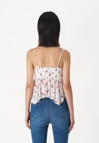 Donna con i capelli neri fino alle spalle, che indossa un top senza maniche bianco a fiori e jeans blu, di spalle rispetto a uno sfondo bianco semplice.