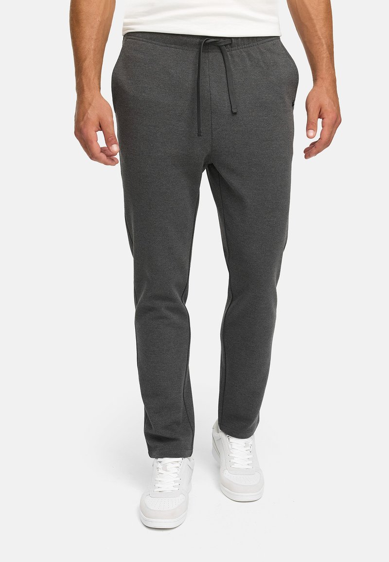 Man wearing dark gray jogger pants with drawstring waist and white sneakers, standing against plain light background.