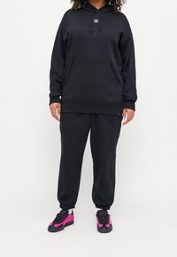 Černá mikina s kapucí, přední kapsou a šňůrkami, v kombinaci s odpovídajícími tepláky. Tenisky s růžovými akcenty a texturovaným povrchem.