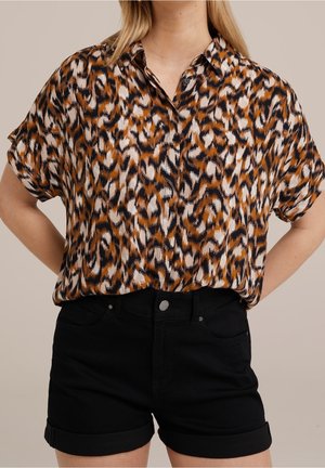 Vrouw die een bruin, zwart en wit patroon korte mouwen blouse draagt, gestopt in zwarte shorts met opgerolde zoom, staand met de handen achter haar rug.