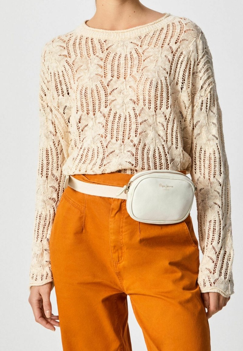 Beige neulepusero, jossa on leikattu kuvio, yhdistettynä oransseihin housuihin. Pienen valkoisen nahkalaukun, jossa on vetoketju, on kiinnitetty vyötärölle.