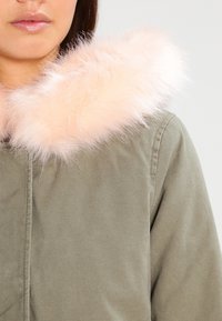 Žena v olivově zeleném kabátu s bledě růžovým načechraným límcem, zkráceným pod nosem a nad pasem, na neutrálním pozadí.