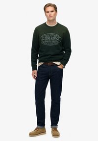 Groene sweatshirt met ronde hals en bedrukte logo, gedragen over een licht shirt. Donkere jeans en beige schoenen maken de look compleet.