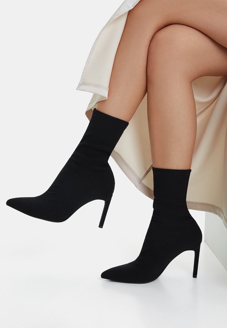 Jambes croisées portant des bottines chaussettes noires à bout pointu avec talons aiguilles et une jupe crème.