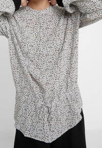 Blouse blanche à motif floral avec manches longues, encolure arrondie et détail froncé à la taille. Tissu doux et léger avec une coupe décontractée.