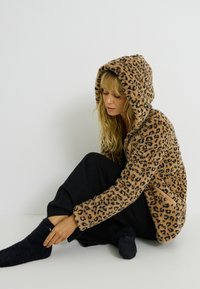 Veste à capuche en molleton doux à imprimé léopard, avec deux poches latérales et fermeture éclair, portée avec un pantalon noir et des chaussettes foncées.