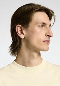 Svetlo bež srajca s kratkimi rokavi in okroglo ovratnikom, prikazana na nevtralni podlagi, ki prikazuje profil modela in njegovo frizuro.