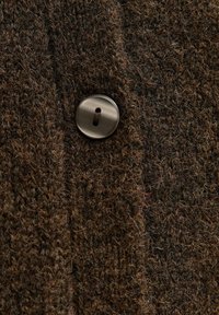 Brązowa tkanina o teksturze, z wyraźnym metalowym guzikiem z dziurką i nitką, typowa dla dzianin lub odzieży wierzchniej.