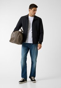 Veste noire légère avec col montant, t-shirt blanc en dessous, jean bleu et baskets noires. Porte un sac de sport en cuir beige.