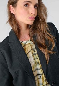 Zwarte krijtstreepblazer over een patroonhemd met groene en gele tinten, met een ronde halslijn en zichtbare knopen.