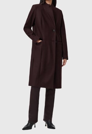 Femme portant un long manteau marron foncé boutonné devant, avec un pantalon et des bottines noires à talons, debout avec une main dans la poche.
