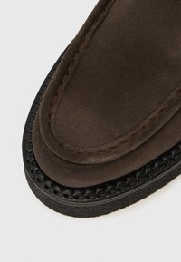 Dunkelbrauner Wildleder-Hausschuh mit rundem Zehenbereich, auffälligen Nähten und einer strukturierten Gummisohle mit gewebten Details an der Kante.