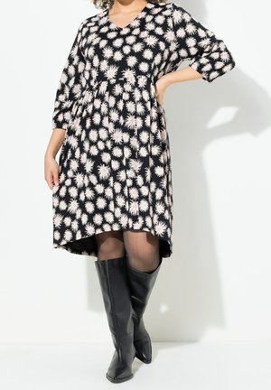 Kobieta w czarnej sukience do kolan z białymi kwiatowymi wzorami, z rękawami do połowy przedramienia, przezroczystych czarnych rajstopach i czarnych kozakach do kolan.