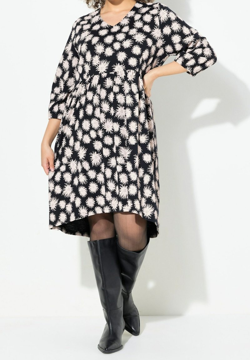Femme portant une robe noire mi-longue à motifs floraux blancs, manches trois-quarts, collants noirs transparents et bottes noires montantes jusqu'aux genoux.