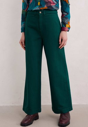 Personne portant un pantalon large taille haute vert foncé, un chemisier multicolore à manches longues et des chaussures marron foncé, debout devant un fond uni.