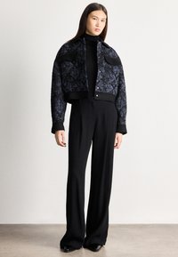 Fiatal nő áll, fekete bőszárú nadrágot, fekete garbót és sötét mintás, kapcsos felsőrészt visel, világos háttér előtt.