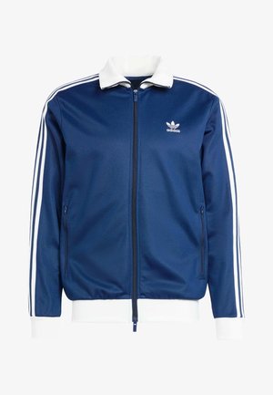 Modra športna jakna z zadrgo in belim ovratnikom ter manšetami, ki ima tri bele črte ob rokavih in logotip Adidas na prsih.