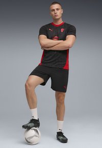 Sort og rød atletisk trøje med korte ærmer, der har et grafisk mønster, parret med sorte shorts og sorte fodboldstøvler, stående på en fodbold.