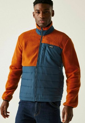 Mężczyzna ubrany w dwukolorową pomarańczową bluzę polarową i niebieską pikowaną kurtkę, stojący z rękami swobodnie opuszczonymi wzdłuż ciała na jednolitym tle.