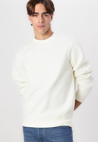 Sudadera blanca de cuello redondo con mangas largas y tela texturizada. Presenta un sutil patrón en la parte delantera. Combinada con unos jeans de mezclilla azules.