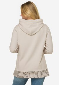 Haut à capuche beige avec une grande capuche texturée et "WEATHER" imprimé en lettres foncées sur l'ourlet arrière. Tissu doux, coupe décontractée et manches longues.
