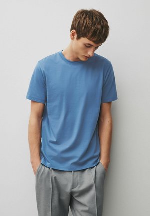 Ung mand iført en blå t-shirt med korte ærmer og grå bukser står med hænderne i lommerne og kigger ned mod en ensfarvet hvid baggrund.