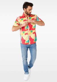 Camicia a maniche corte con un motivo astratto multicolore caratterizzato da cerchi rossi, gialli, verdi e blu, abbinata a jeans blu chiaro e sneakers bianche.