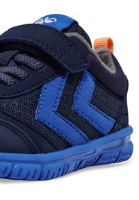 Tamsiai mėlyni sportiniai bateliai su tekstūruotu viršumi, išryškinančiais mėlynos spalvos zigzago akcentais, Velcro užsegimu ir oranžiniu kilimėliu ant kulno.