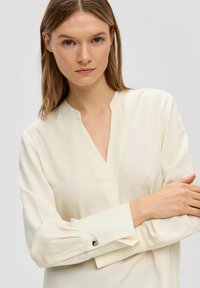 Cremfarbene Bluse mit V-Ausschnitt, langen Ärmeln und geknöpften Manschetten. Glatte Textur mit einem subtilen Glanz, die minimalistische Designelemente aufweist.