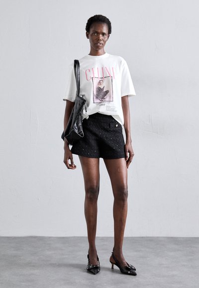 T-shirt graphique blanc avec lettrage rose et image, associé à un short texturé noir avec un bouton doré. Sac noir et talons.