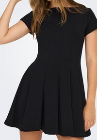 Femme portant une robe courte noire cintrée avec des manches courtes et des plis, montrée du cou jusqu'à mi-cuisse, sur un fond uni clair.