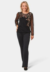 Schwarzes Oberteil mit einem paillettenbesetzten Blumenmuster und transparenten langen Ärmeln. Kombiniert mit dunklen, funkelnden geraden Hosen und schwarzen Schuhen.